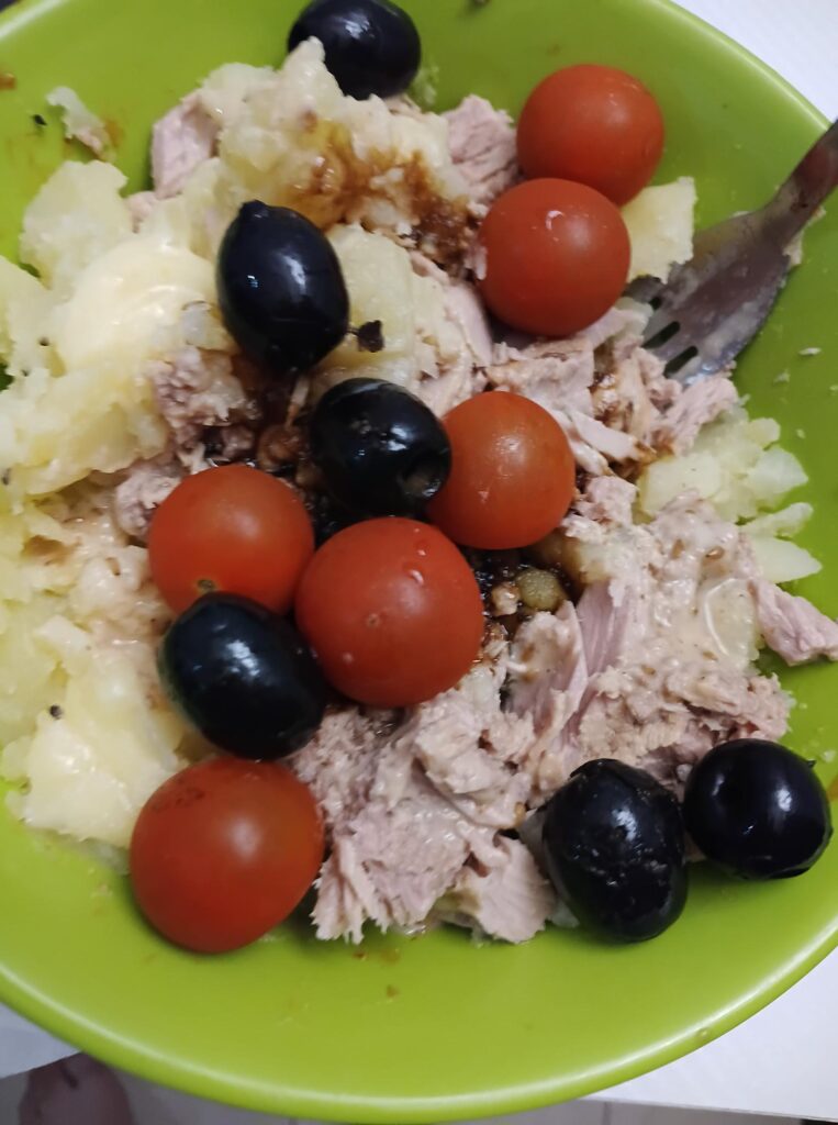 Une salade composée de pommes de terre, thon, olives noires et tomates cerises dans un bol vert avec une fourchette en métal.