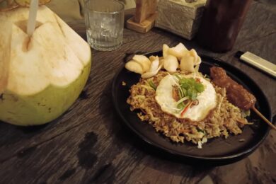 Une assiette de riz frit indonésien surmontée d'un œuf au plat, accompagnée d'une brochette de poulet et d'une noix de coco fraîche ouverte sur une table en bois sombre.