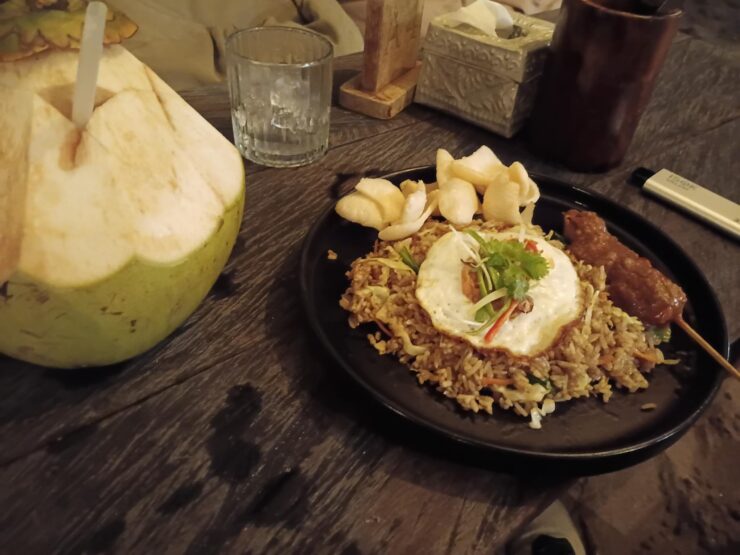 Une assiette de riz frit indonésien surmontée d'un œuf au plat, accompagnée d'une brochette de poulet et d'une noix de coco fraîche ouverte sur une table en bois sombre.