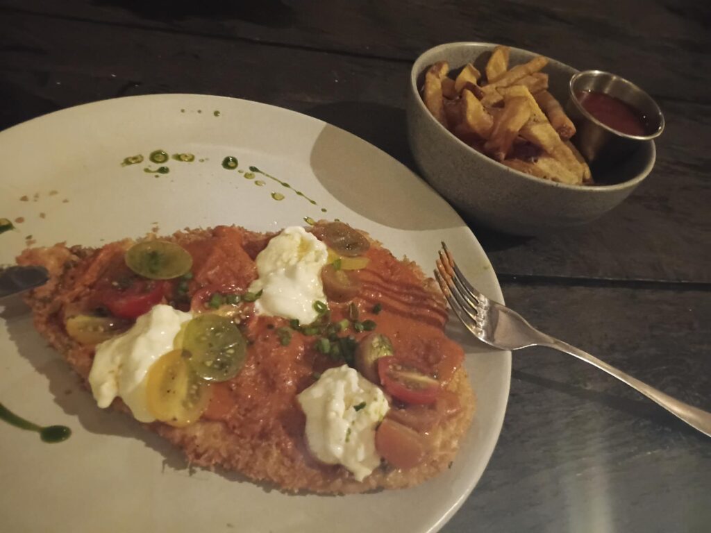 Un plat de poulet pané recouvert de sauce tomate, mozzarella et tomates cerises, servi avec un bol de frites et de la sauce ketchup dans un restaurant de type brasserie.