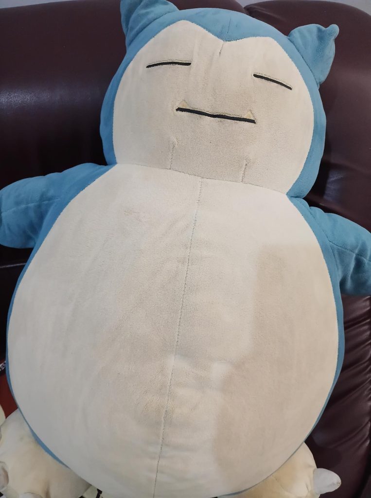 Une imposante peluche de Ronflex (Snorlax) endormi, avec sa fourrure bleue et son gros ventre beige, installée sur un canapé en cuir marron.
