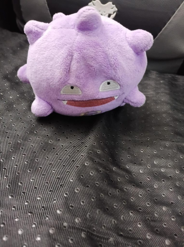Gros plan sur une peluche violette de Smogo (Koffing) avec les yeux et le sourire niais caractéristiques de Métamorph (Ditto), posée sur un tissu noir perforé. Cette peluche originale, illustre les goodies uniques que l'on trouve uniquement au Japon.