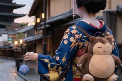 Vue de dos d'une jeune femme en kimono bleu marine orné de motifs Pikachu et d'éclairs dorés, déambulant dans une rue pavée traditionnelle de Kyoto au crépuscule. Elle porte un grand sac à dos en peluche Ronflex (Snorlax) et tient une petite breloque Smogo (Koffing) à la main. Sa coiffure traditionnelle est surmontée d'accessoires en forme d'oreilles de Pikachu, avec la pagode Yasaka en arrière-plan.