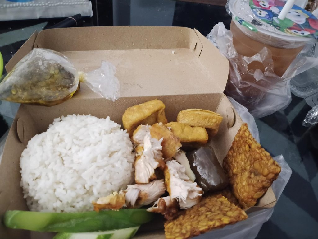Un repas à emporter dans une boîte en carton contenant du riz blanc, du poulet frit, du tofu, du tempeh, des morceaux de concombre et un sachet en plastique rempli de sambal vert, accompagné d'une boisson dans un gobelet en plastique.