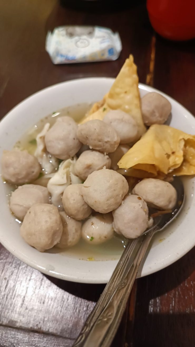 Un bol de soupe Bakso rempli de boulettes de viande rondes et de morceaux de wonton frits croustillants, avec une cuillère posée à l'intérieur.