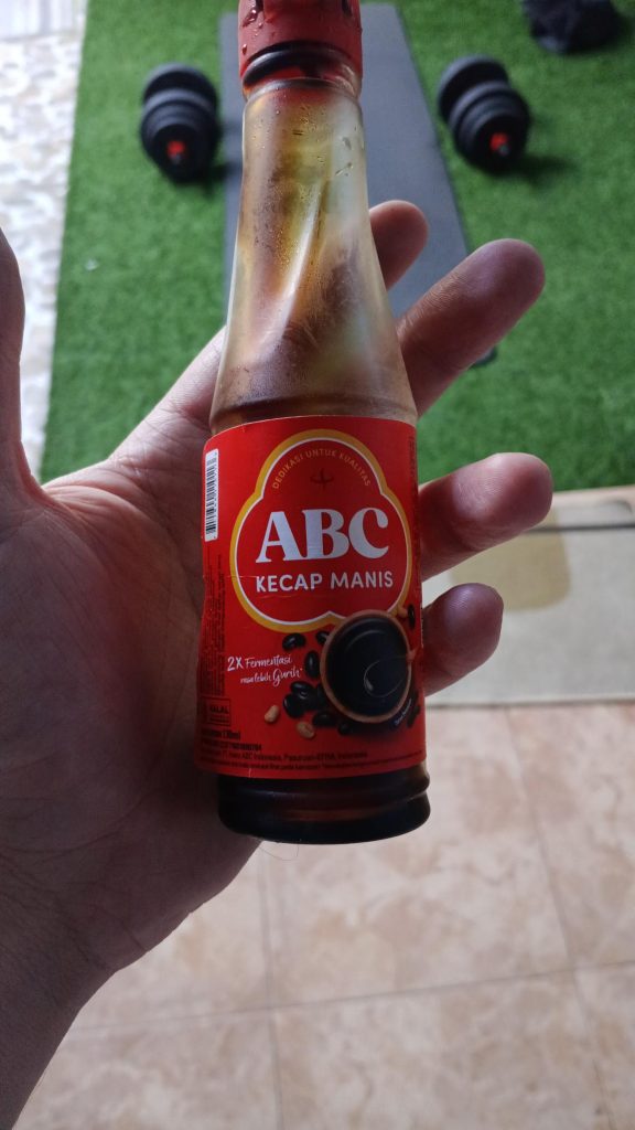Une main tient une bouteille de sauce soja sucrée de la marque ABC (Kecap Manis) avec une étiquette rouge distinctive, au-dessus d'une surface qui semble être un tapis de sport.