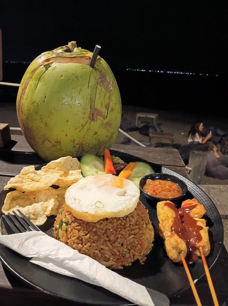 Une assiette de Nasi Goreng avec un œuf au plat sur le dessus, accompagnée de brochettes de satay, de chips de crevettes (krupuk), de tranches de concombre et d'une petite portion de sambal, avec une noix de coco entière en arrière-plan.