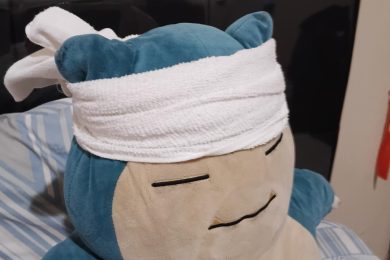 Un grand peluche Snorlax est allongé sur un lit à rayures, avec une serviette blanche nouée autour de sa tête, donnant l’impression qu’il se repose ou qu’il récupère.