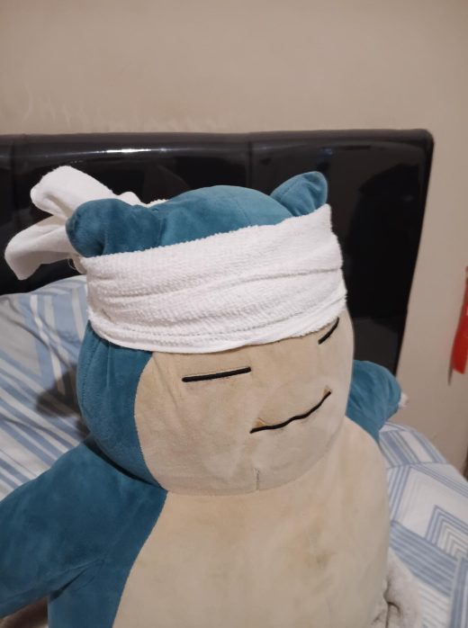Un grand peluche Snorlax est allongé sur un lit à rayures, avec une serviette blanche nouée autour de sa tête, donnant l’impression qu’il se repose ou qu’il récupère.