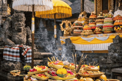 Temple hindou balinais avec offrandes colorées de fleurs, fruits et encens disposées sur un autel en pierre, entouré de parasols cérémoniels et de sculptures traditionnelles.